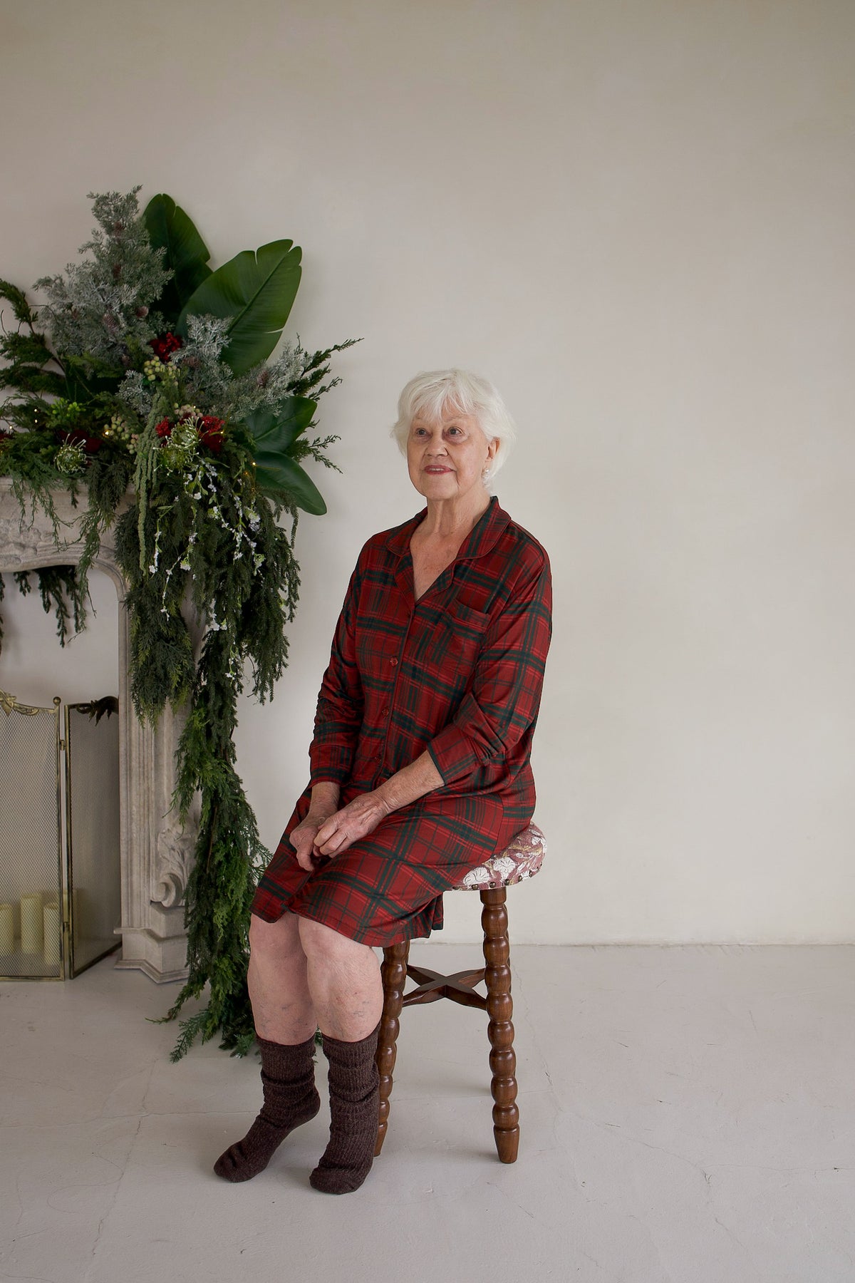 An older woman with white hair wears a Women’s Bamboo Sleeping Dress | Christmas Plaid and brown knit socks while sitting on a small wooden stool.