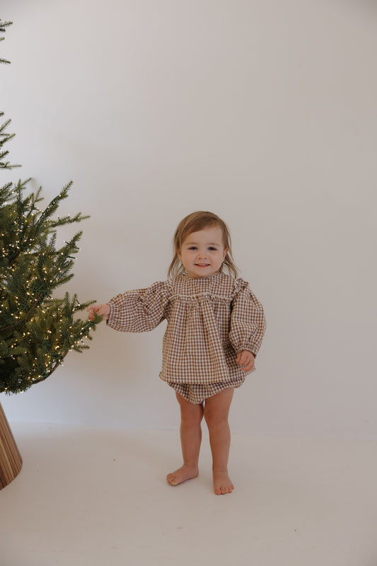 A smiling toddler stands barefoot indoors by a lit Christmas tree, wearing the Bloomer Set | Honey Gingham from forever french baby. The cozy set features long sleeves and shorts. The background is plain white.