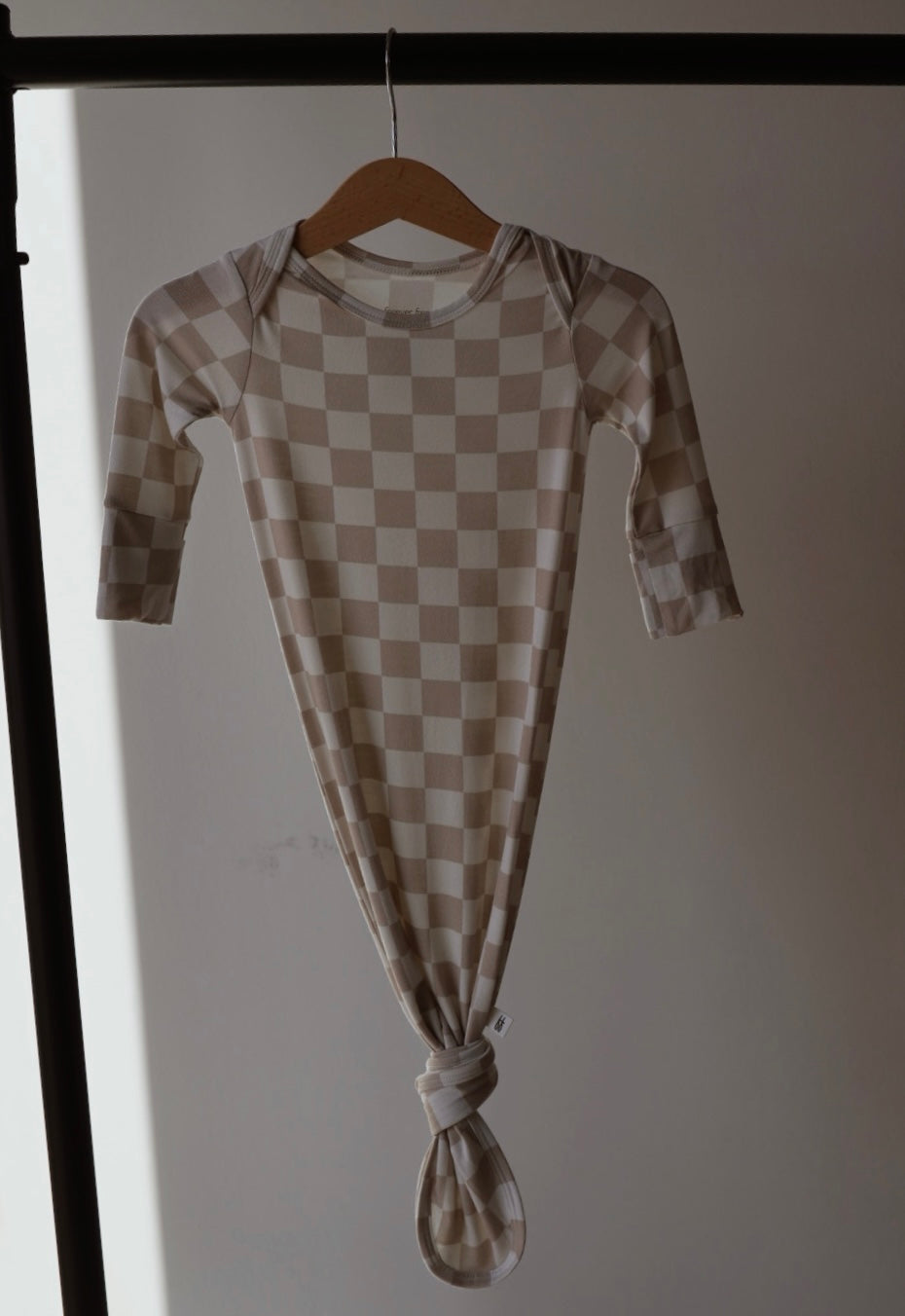The Bamboo Knotted Gown | Checkerboard by forever french baby features a beige and cream checkered design with long sleeves—stylish and soft for newborns, shown hanging on a wooden hanger with sunlight in the background.