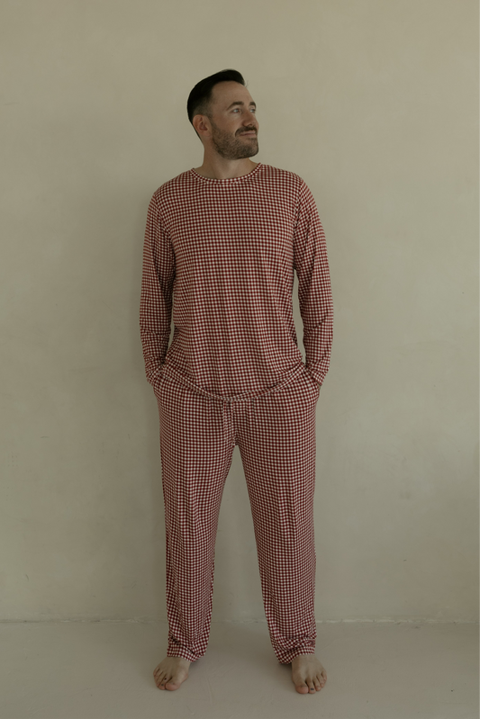 A man stands barefoot by a beige wall, smiling slightly, wearing forever french baby Men's Bamboo Pajamas in Red Gingham with his hands in his pockets.