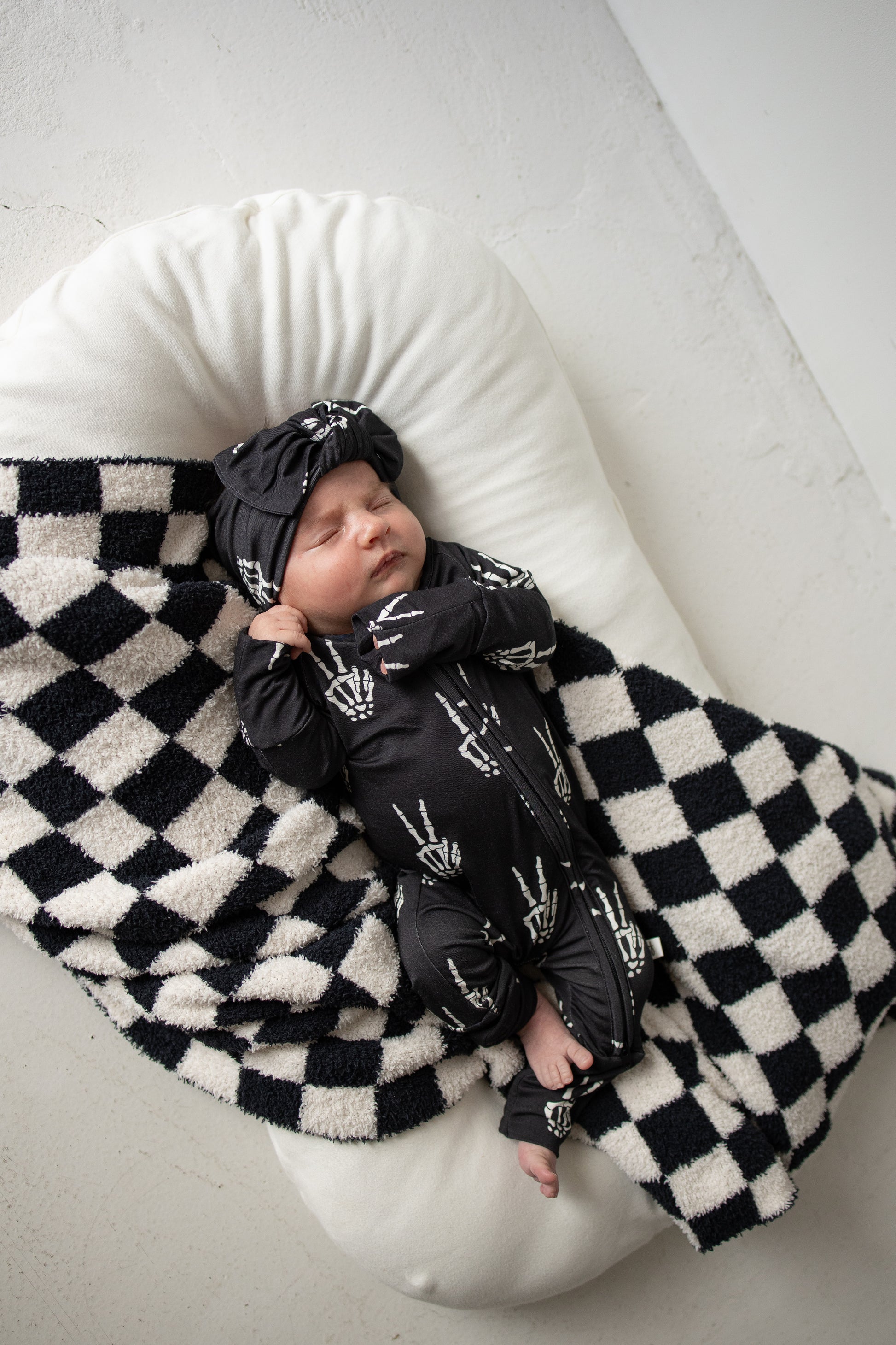 A baby in forever french baby’s Bamboo Zip Pajamas | Midnight Skelly and matching hat sleeps peacefully on a white cushion, partially covered with a black and white checkered blanket. These hypo-allergenic pajamas offer comfort and safety for your little one’s delicate skin.