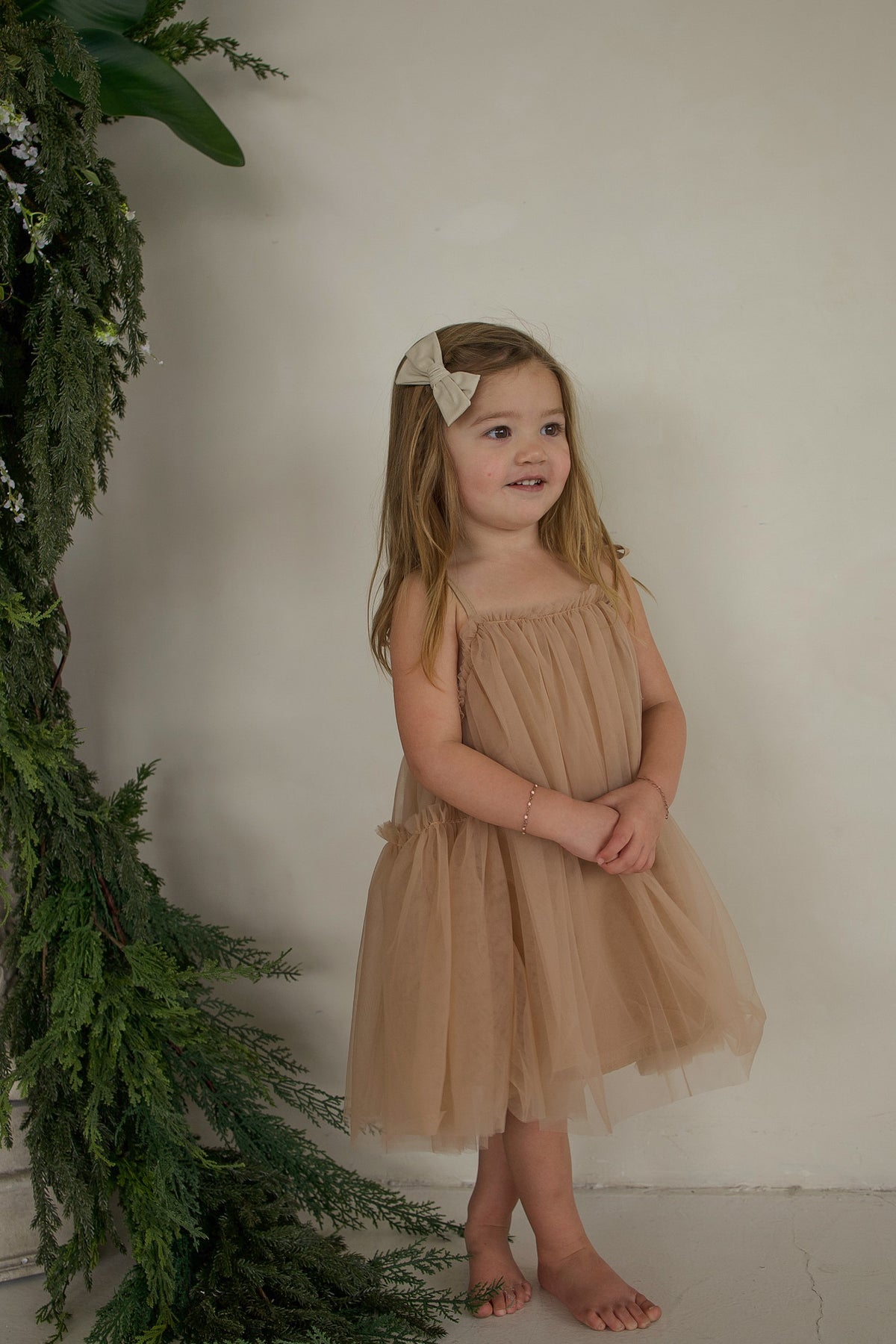 A young girl wearing a festive, beige Tutu Dress | Teddy with matching tulle bow straps and a hair bow.