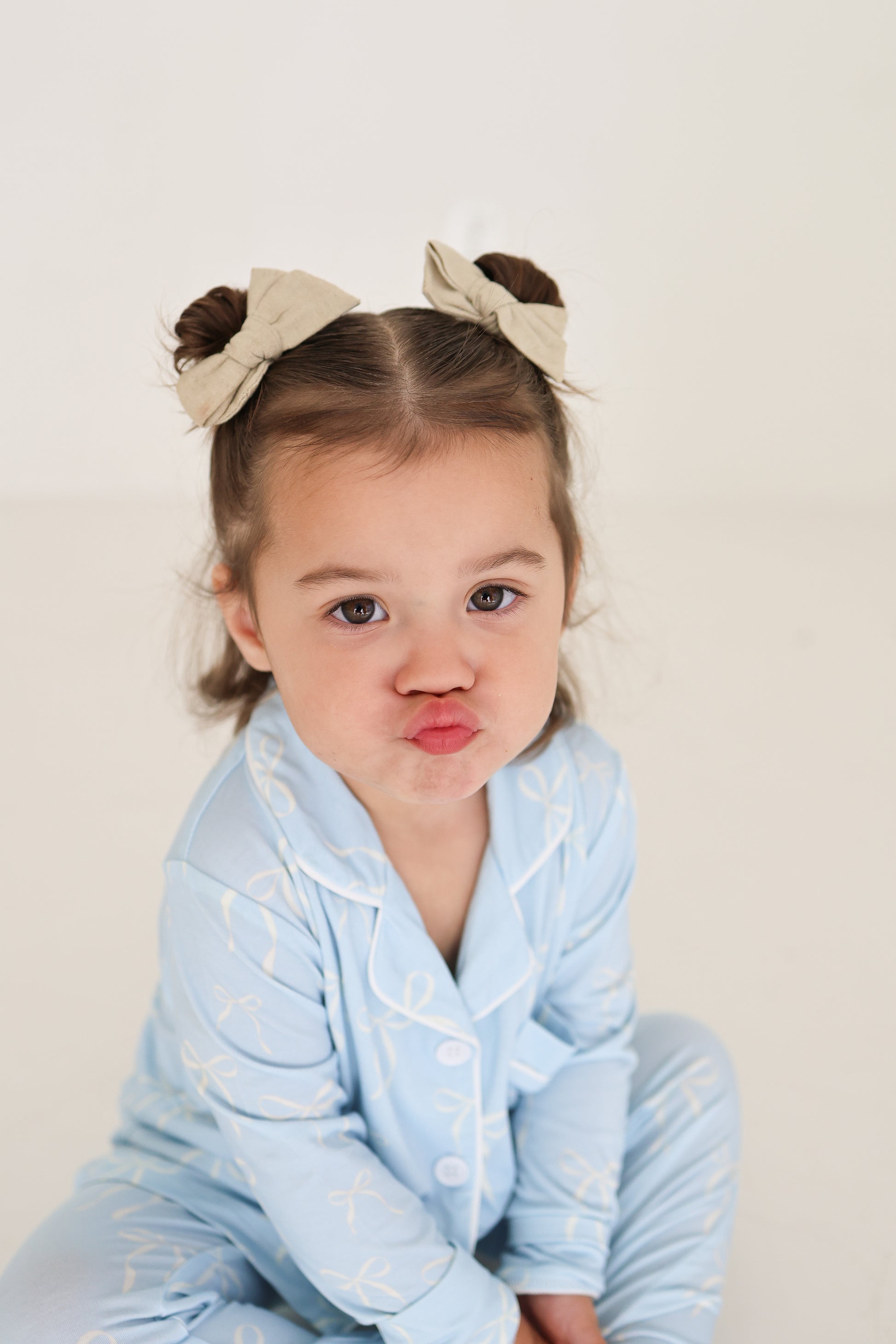 A young girl with brown hair in two buns and beige bows poses playfully on the floor, wearing the forever french baby Girl’s Bamboo Button Up Lounge Set | Blue Bows. The background is plain and light-colored.