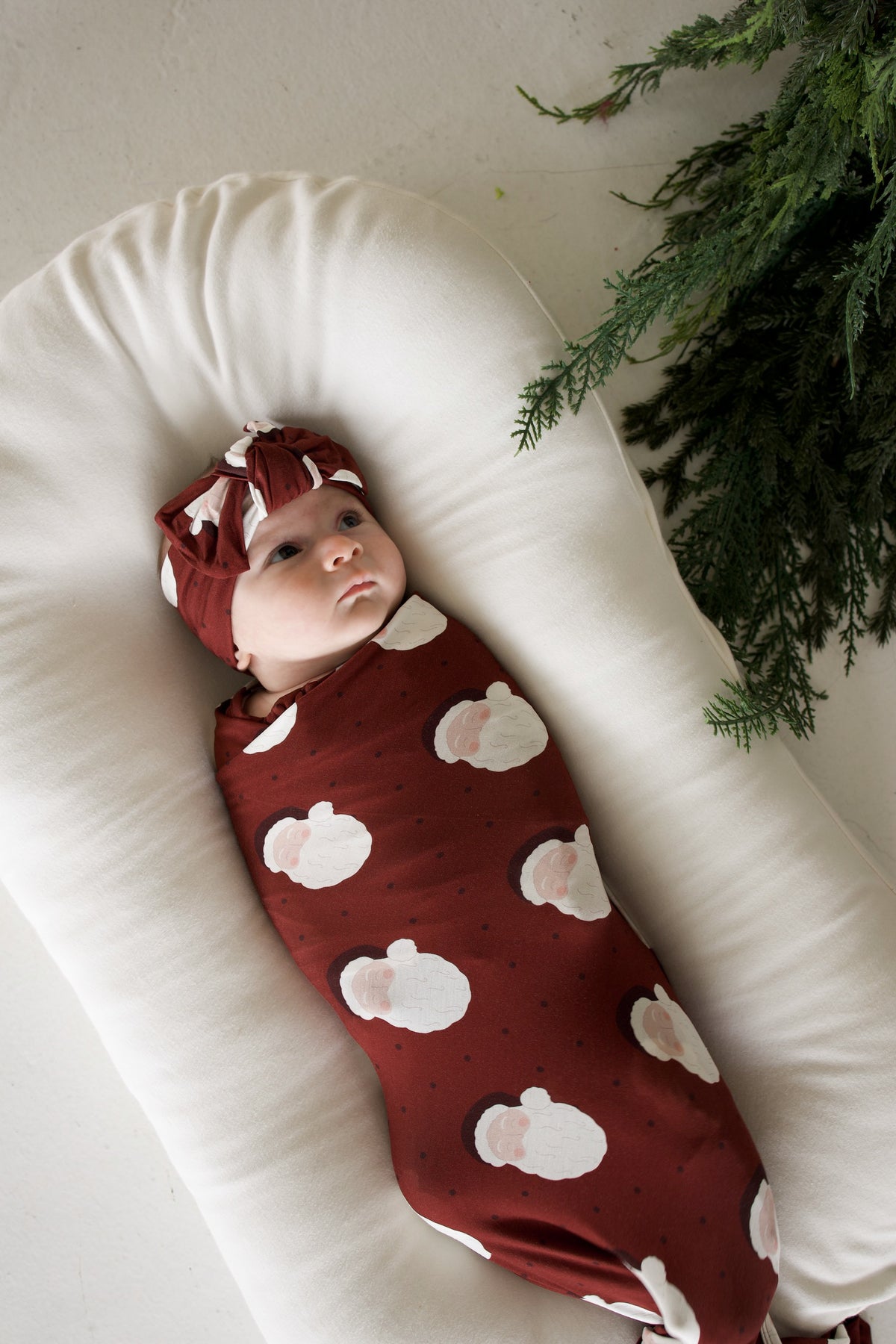 A baby wearing a matching headband and a Bamboo Swaddle | Vintage Santa, resting in a cream-colored lounger.