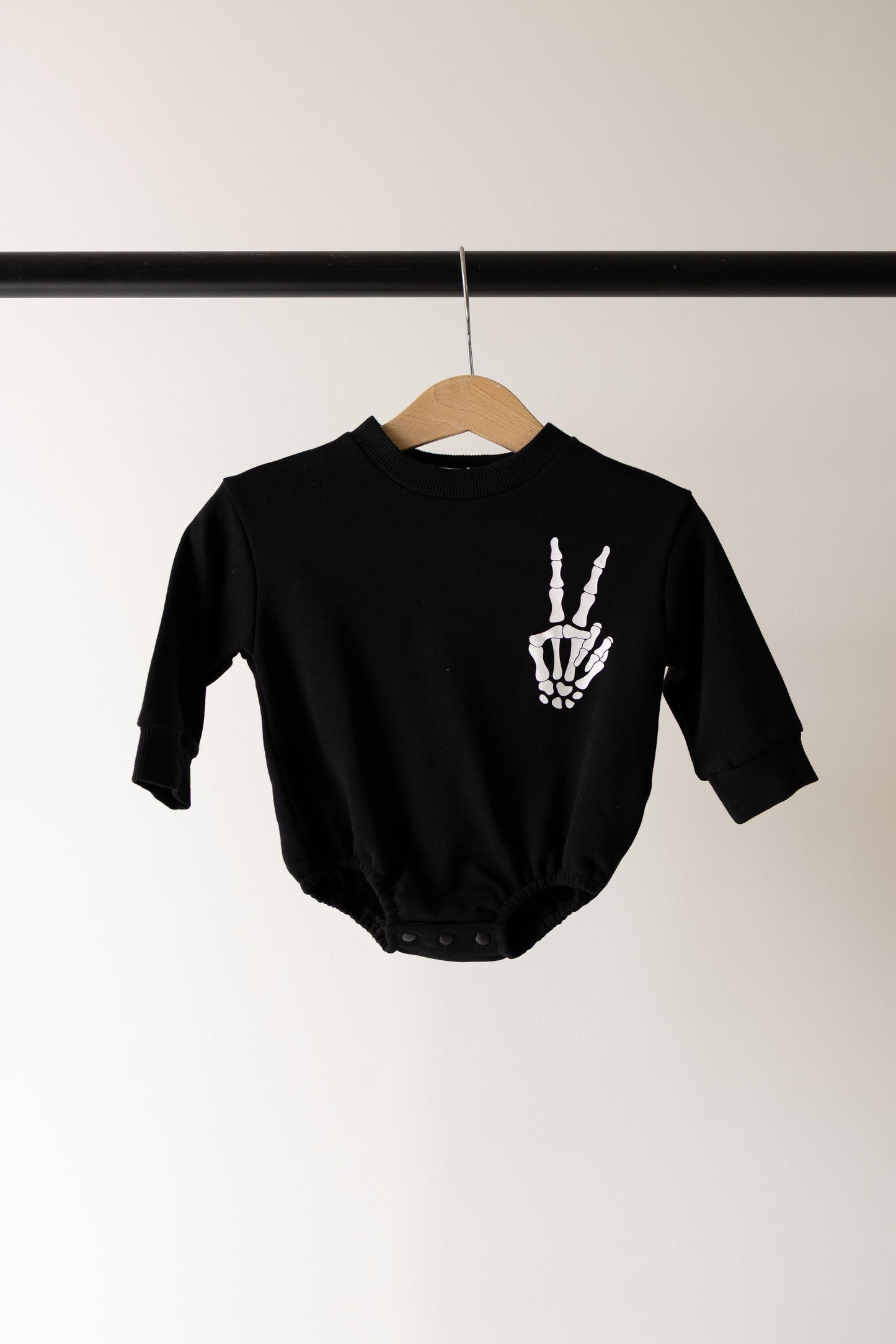 A black Sweatshirt Romper from forever french baby, named Midnight Skelly, hangs on a wooden hanger. It features a white skeleton hand making a peace sign on the chest and is crafted from soft newborn cotton material. Inspired by playful designs and offering both comfort and spooky charm, this cozy romper is perfect for your little one.