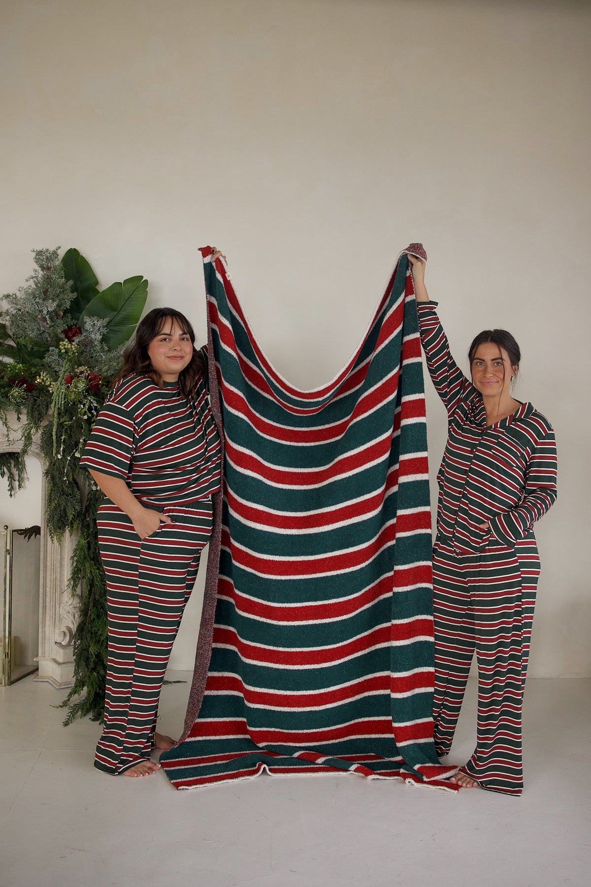 Two women in matching holiday-striped pajamas hold up a large, red, green, and white Holiday Stripe Plush Blanket.