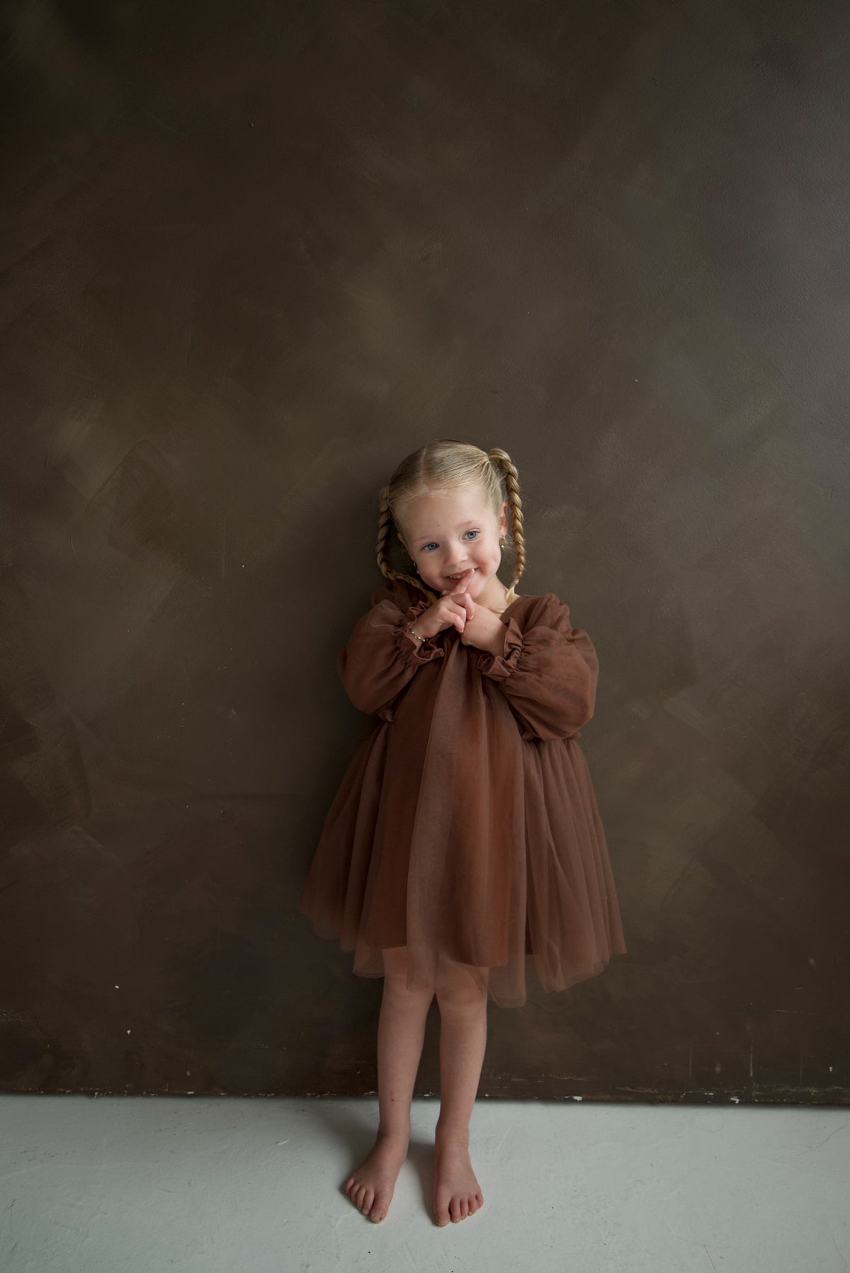 A young girl wearing a cocoa Long Sleeve Tutu Dress with bow detail stands against a matching brown wall.