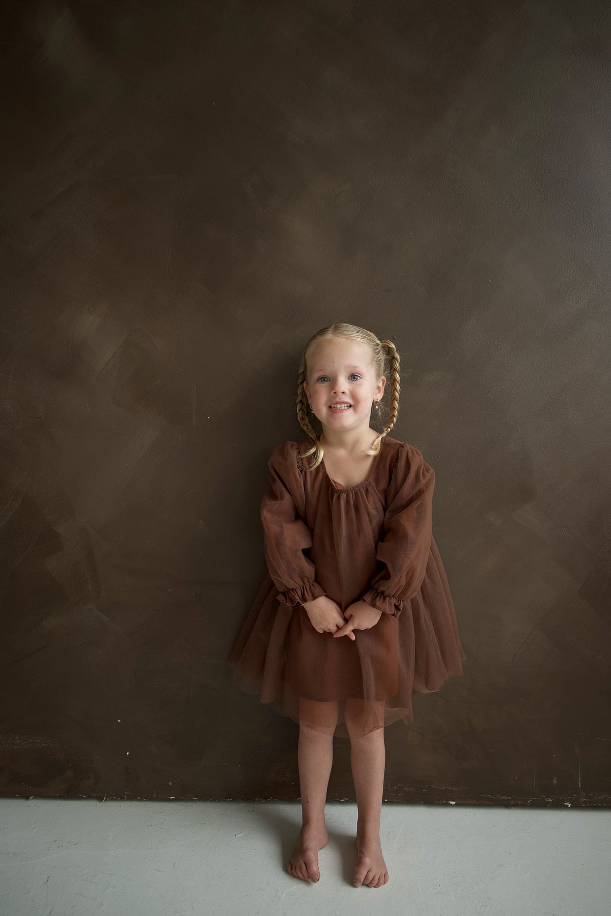 A young girl stands against a brown wall wearing a Long Sleeve Tutu Dress | Cocoa, a festive long-sleeve dress with a gathered skirt and bow detail.
