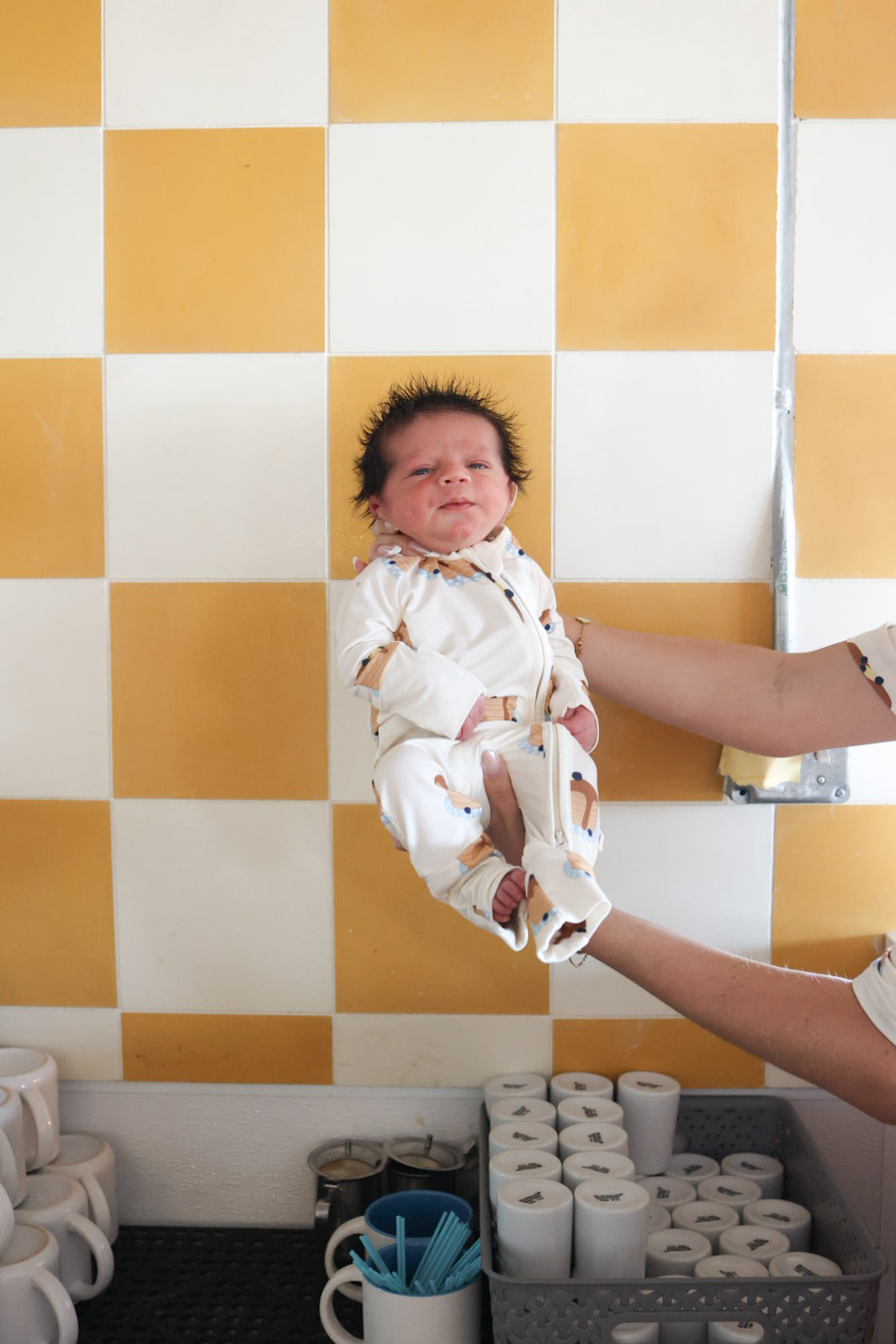 A baby in forever french baby Bamboo Zip Pajamas | Sunday Stack is held up by an adult's hand in front of a yellow and white checkered wall, above a counter with stacked white cups and containers.