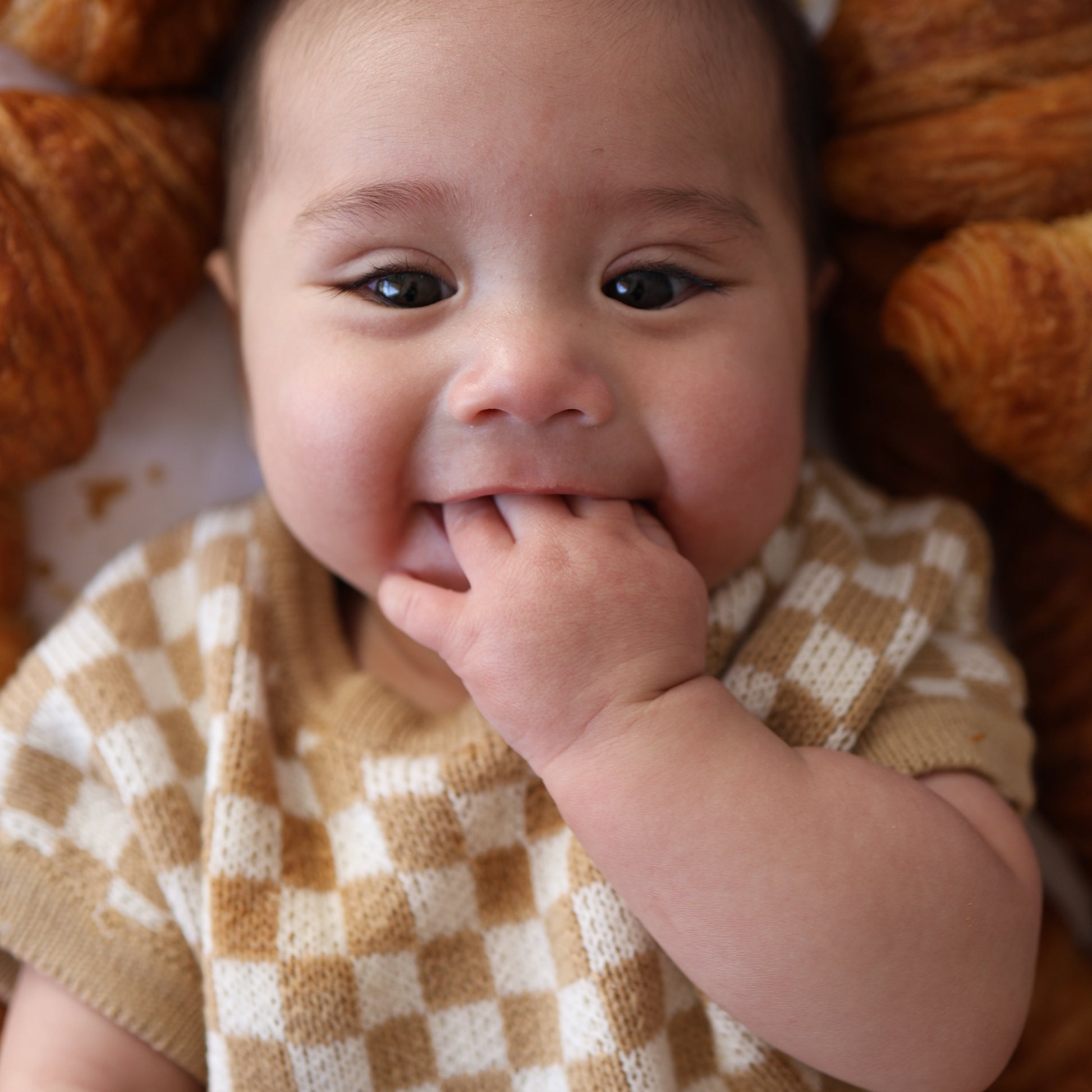 A baby in a forever french baby Knit Romper | Latte Check with beige and white checks lies among croissants, smiling and sucking on their fingers.