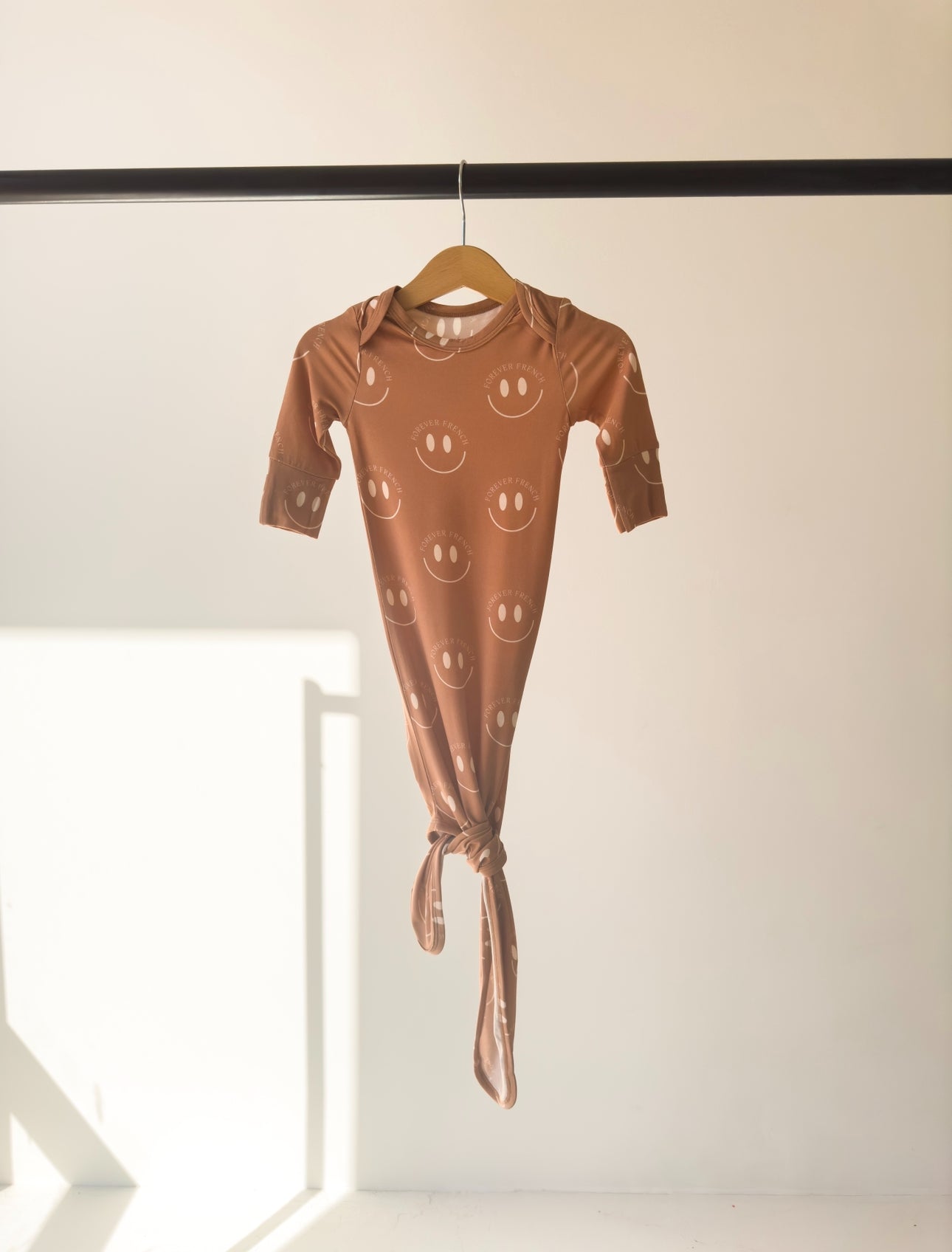 A Bamboo Knotted Gown | Desert Clay ff Smile by forever french baby, hangs from a wooden hanger on a horizontal rod. Sunlight casts warm shadows on the wall and floor, highlighting the hypo-allergenic bamboo garment adorned with smiley faces in a minimalistic scene.