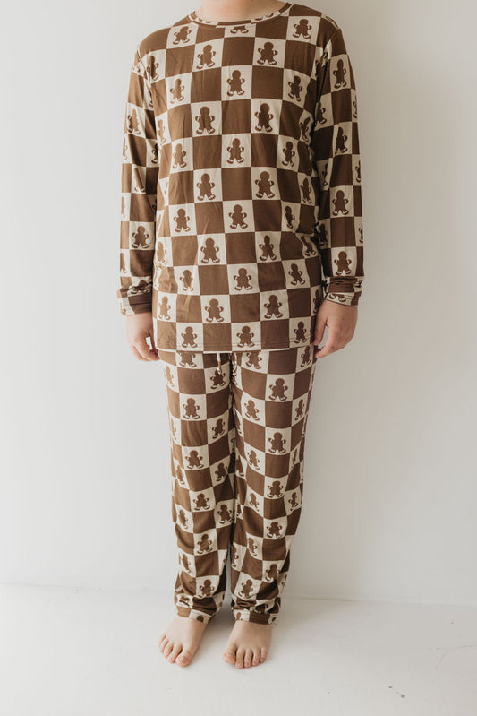 A person wearing forever french baby's Teen Bamboo Pajamas in the Gingerbread Checkerboard pattern stands barefoot against a plain white background. These breathable, long-sleeved pajamas envelop the body in hypo-allergenic comfort, ideal for a peaceful night's sleep.