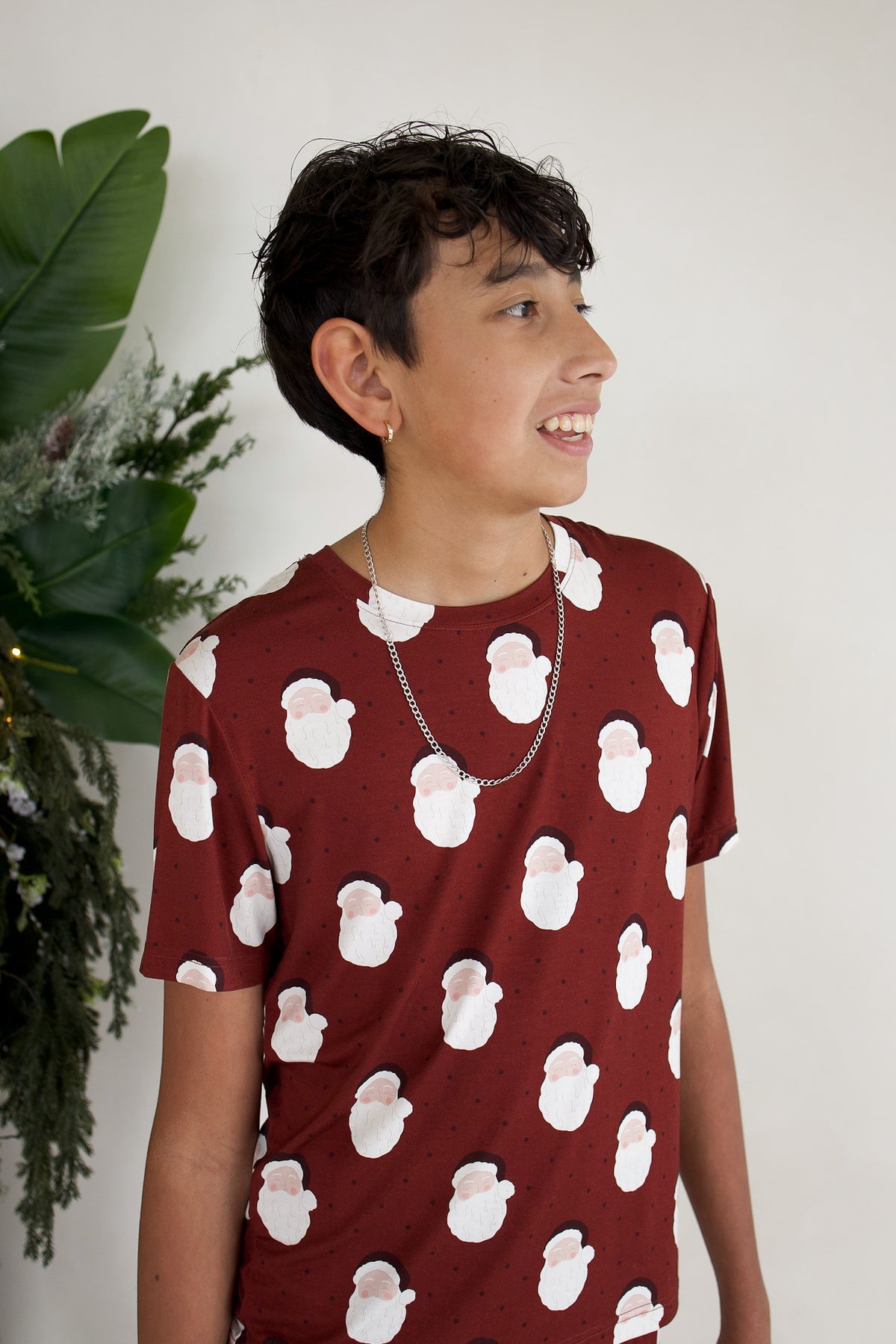 A child wears Youth Straight Leg Bamboo Pajamas | Vintage Santa, consisting of a maroon short-sleeved shirt patterned with small Santa Claus faces.