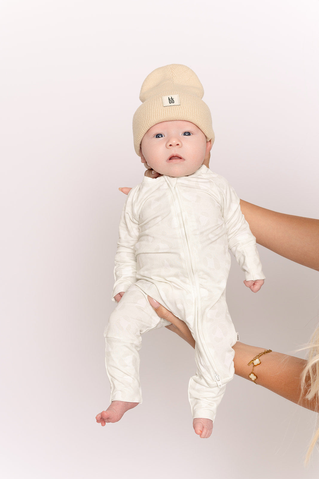 A baby in forever french baby Bamboo Zip Pajamas | Meow and a beige knit beanie is held upright by an adult with a gold bracelet, set against a plain light background.