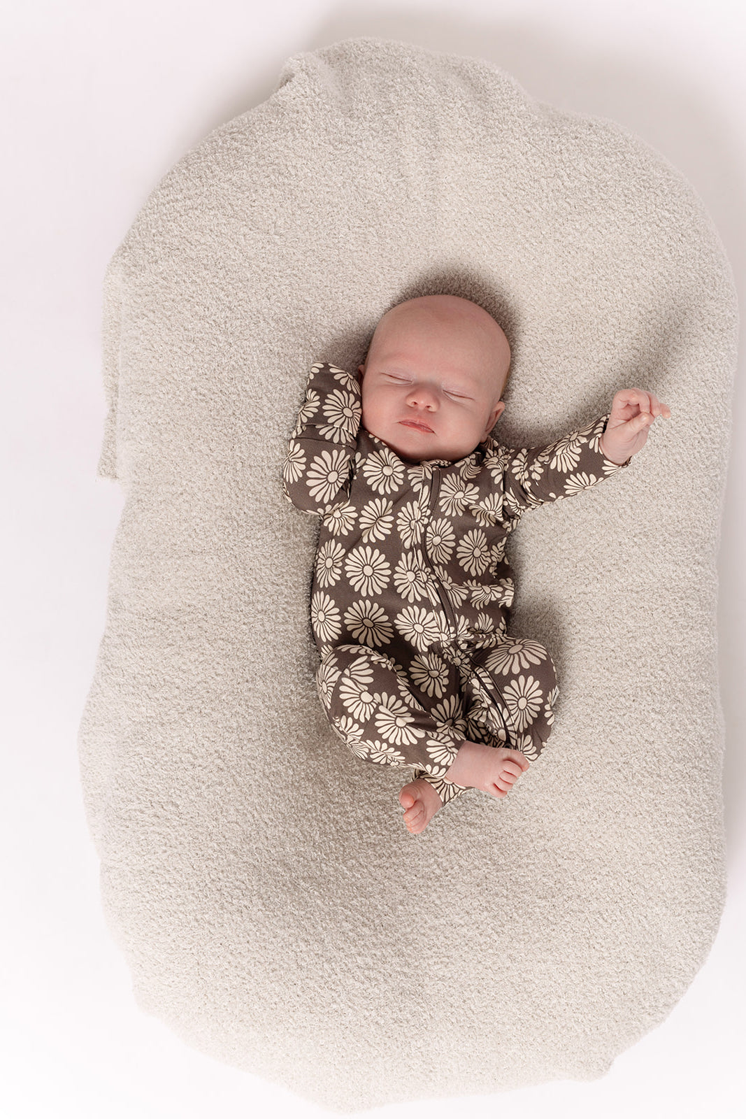 A sleeping baby rests on a textured cream blanket, wearing forever french baby's Bamboo Zip Pajamas in Retro Floral—a breathable brown onesie with white daisies—arms raised in comfort for a peaceful night's sleep.