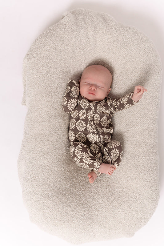 A sleeping baby rests on a textured cream blanket, wearing forever french baby's Bamboo Zip Pajamas in Retro Floral—a breathable brown onesie with white daisies—arms raised in comfort for a peaceful night's sleep.