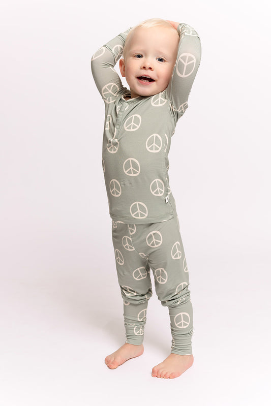 A barefoot toddler with blonde hair smiles and stretches in forever french baby Bamboo Two Piece Pajamas | Peace, featuring matching sage green fabric and large white peace sign prints—perfect kids sleepwear. White background.