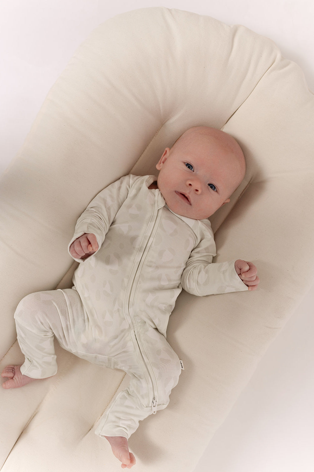 A baby in forever french baby's Bamboo Zip Pajamas | Meow—breathable, hypoallergenic, cream-colored onesie with a leaf pattern—relaxes on a soft cream pad, gazing upward with hands gently raised.