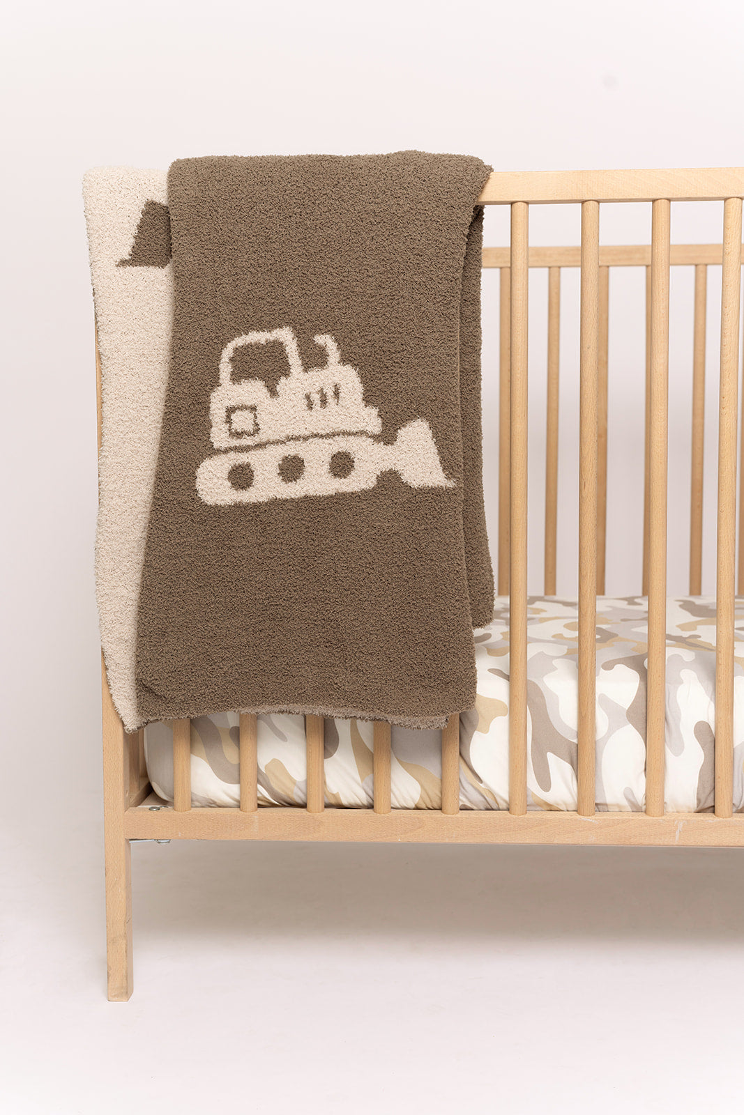 A wooden crib with a beige and white camo-patterned mattress is complemented by the forever french baby Plush Blanket | Bulldozer, a soft microfiber blanket with a bulldozer design draped over the side for comfort and style.