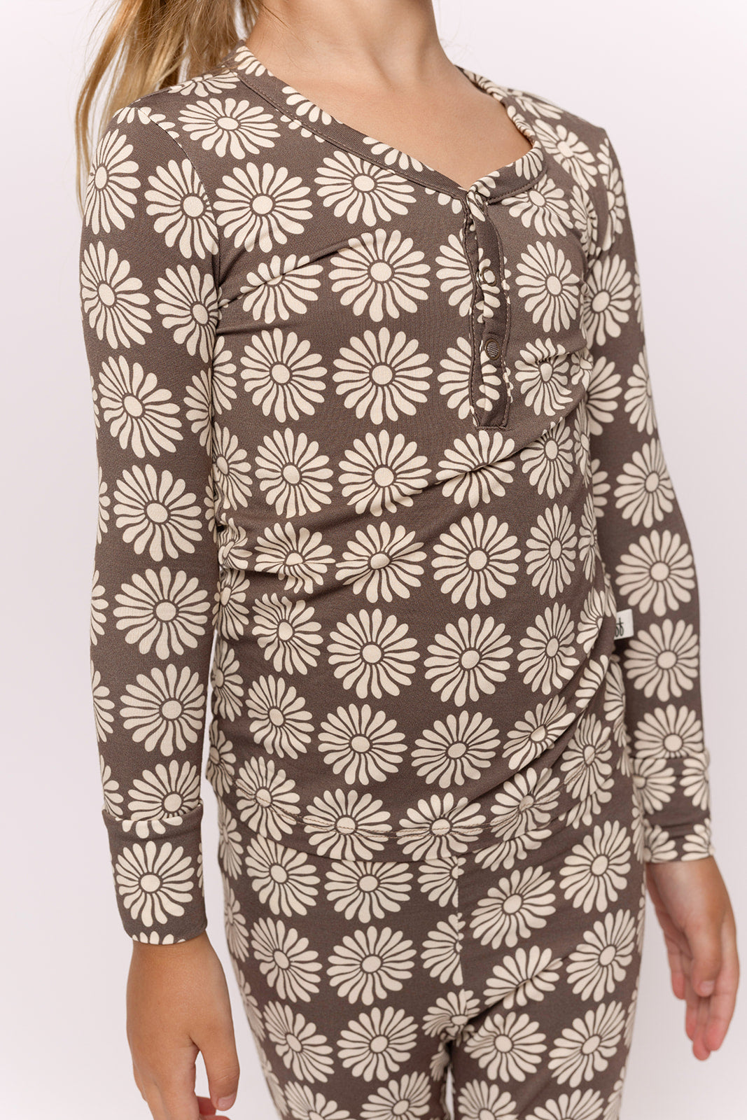 A child models the forever french baby Bamboo Two Piece Pajamas in Retro Floral—a brown long-sleeve set with large white daisies—against a plain light background. The child's head is cropped out of the image.