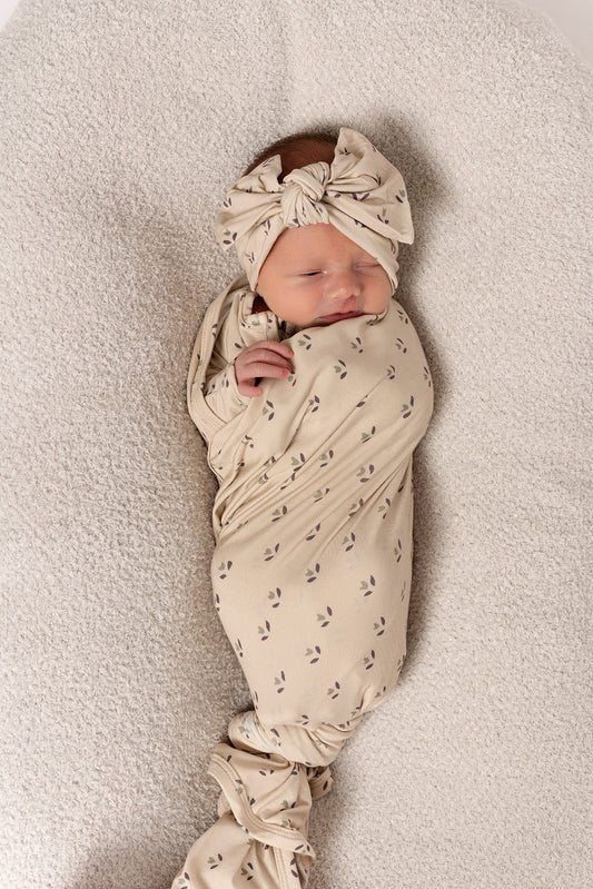 A newborn is wrapped in the forever french baby Bamboo Swaddle | Flora, featuring a breathable fabric with a small leaf pattern and a matching bow headband, resting on a soft, textured surface.