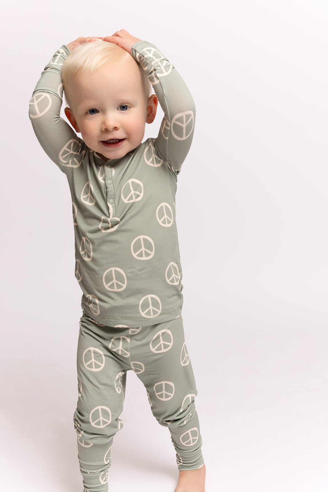 A toddler with blonde hair stands barefoot on a light background, wearing forever french baby's Bamboo Two Piece Pajamas | Peace—hypo-allergenic kids sleepwear with a peace sign print—arms raised overhead, smiling gently.