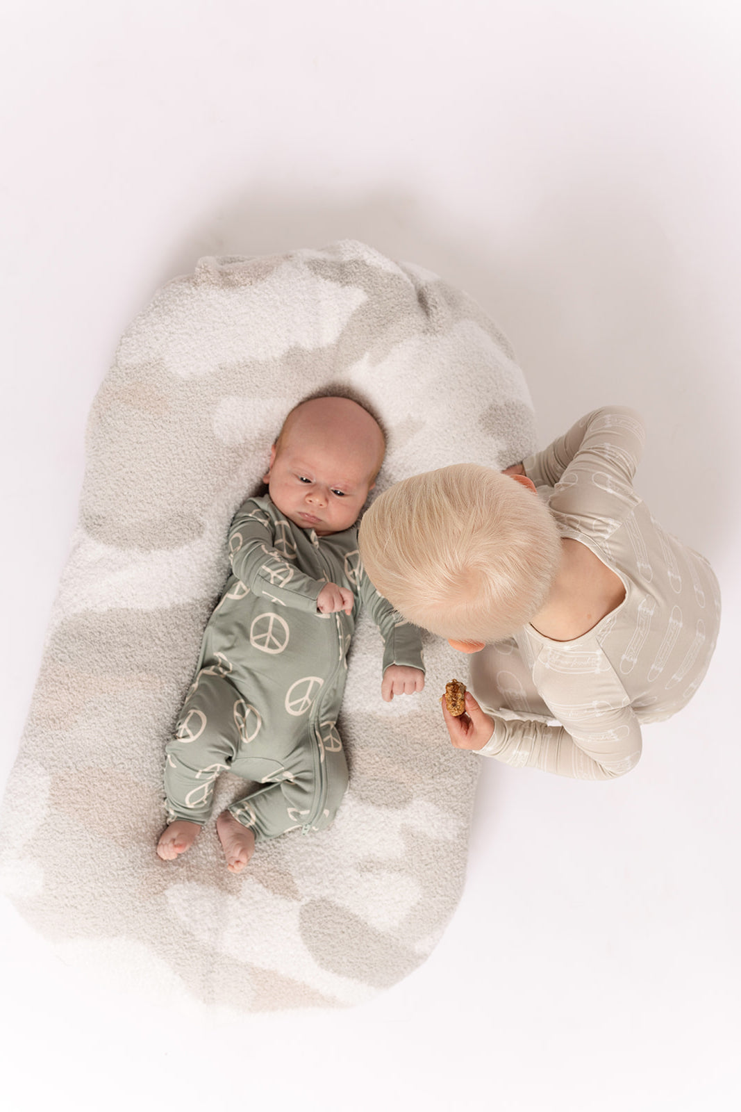 A baby in breathable, hypoallergenic Bamboo Zip Pajamas | Peace by forever french baby lies on a soft cushion, while a blond toddler in cozy bamboo PJs sits nearby holding a cookie. The background is white.