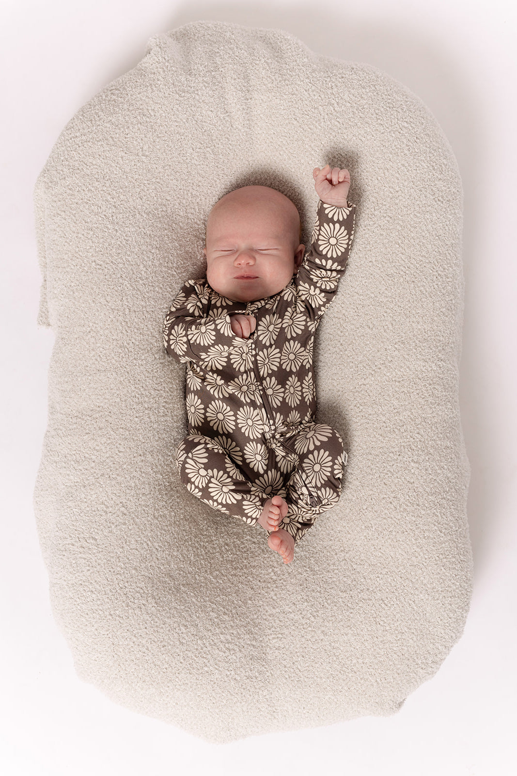 A sleeping baby in forever french baby Bamboo Zip Pajamas | Retro Floral lies on a soft, light cushion, one arm raised and the other by their side—super comfy hypoallergenic onesie perfect for sensitive skin.