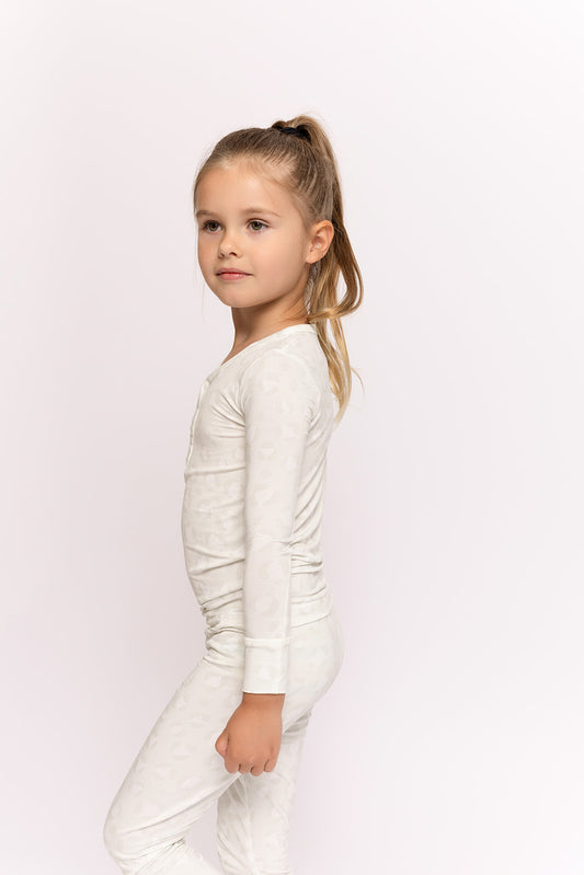 A young girl with long, light brown hair in a ponytail stands sideways, wearing the forever french baby Bamboo Two Piece Pajamas | Meow. The light-colored, breathable set features a long-sleeve top and pants for all-day comfort against a white background.