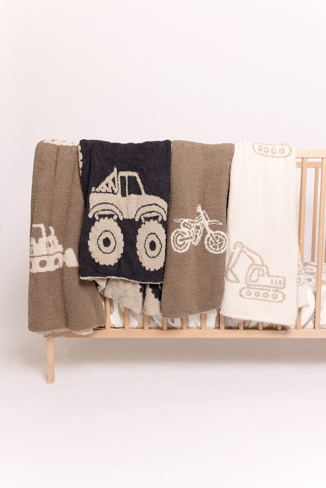 The forever french baby Plush Blanket | Dirt Bike, Oeko-Tex certified, showcases soft microfiber with dump truck, monster truck, and dirt bike prints in neutral tones, displayed on a light wooden crib against a white background.