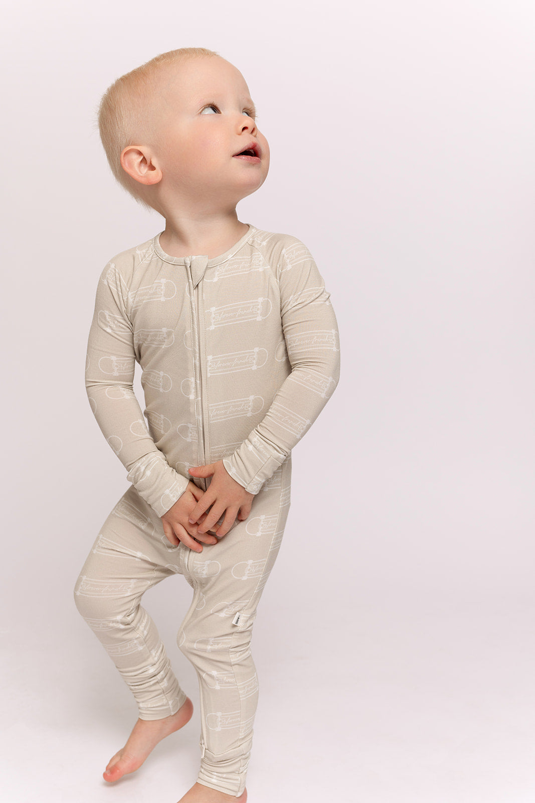 A toddler with short blond hair wears forever french baby's Bamboo Zip Pajamas | Sk8er Boy—beige, long-sleeve, zip-up pajamas with a safety pin print. Barefoot on a light background, they showcase this comfy, hypoallergenic baby outfit.