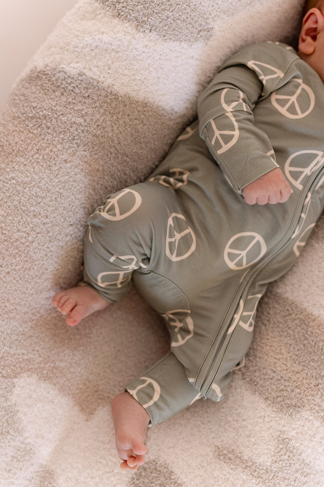 A baby in forever french baby's Bamboo Zip Pajamas | Peace—a green onesie with white peace signs—rests on a striped blanket, with only the lower face, arms, and legs showing.