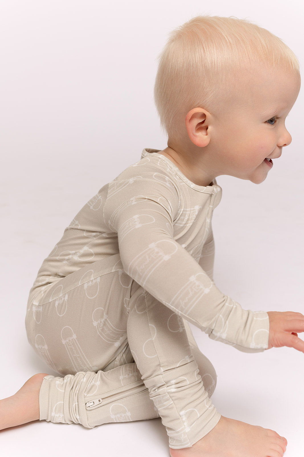 A smiling toddler with light blonde hair sits on the floor in forever french baby's Bamboo Zip Pajamas | Sk8er Boy—a breathable, long-sleeve beige onesie with a white safety pin pattern designed for all-day comfort.