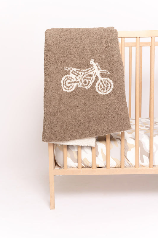 A tan crib features a camouflage bedding set and the forever french baby Plush Blanket | Dirt Bike—a brown microfiber blanket with a white dirt bike outline—draped over the side.