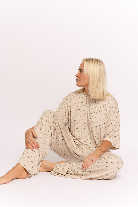 A woman with straight blonde hair sits barefoot on the floor, wearing the forever french baby Women's Boxy Tee Lounge Set | Flora, featuring a loose beige fit with a black pattern, highlighting stylish hypo-allergenic comfort against a white background.
