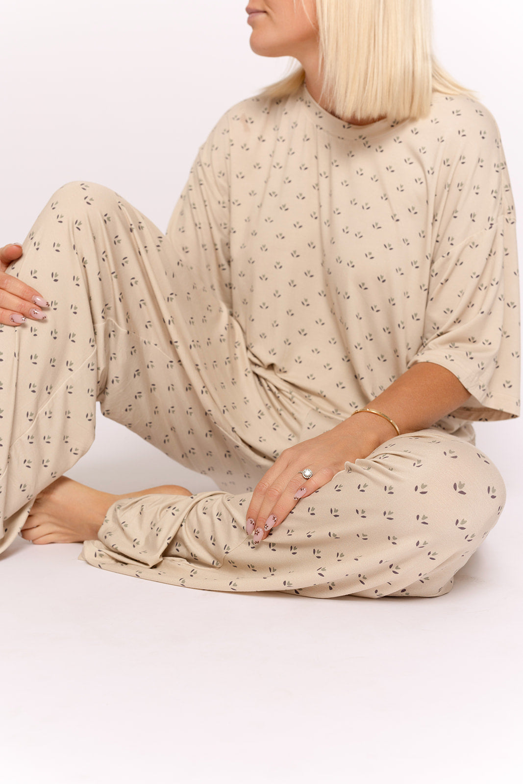 A woman with straight blonde hair sits cross-legged in the forever french baby Women's Boxy Tee Lounge Set | Flora, a loose beige bamboo set with a small black pattern. She wears rings and a bracelet, facing slightly away against a plain white background.