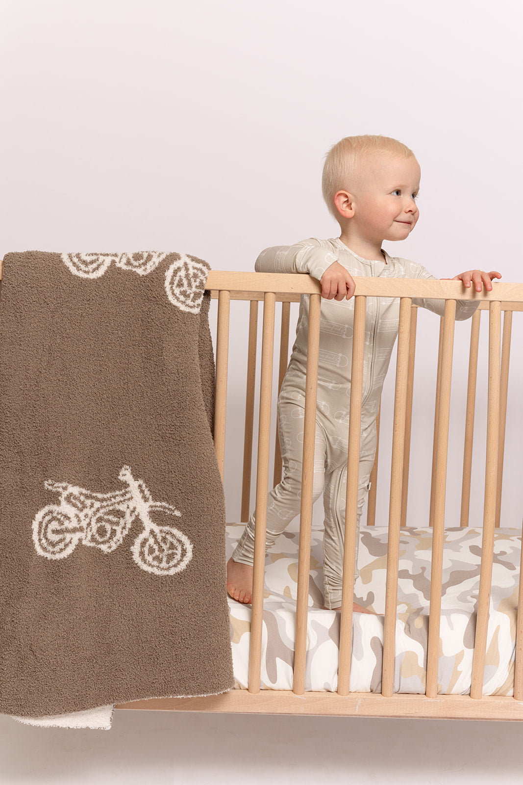 A toddler in light pajamas stands in a wooden crib, with the forever french baby Plush Blanket | Dirt Bike draped over the side; the crib mattress features a white and beige camouflage pattern.