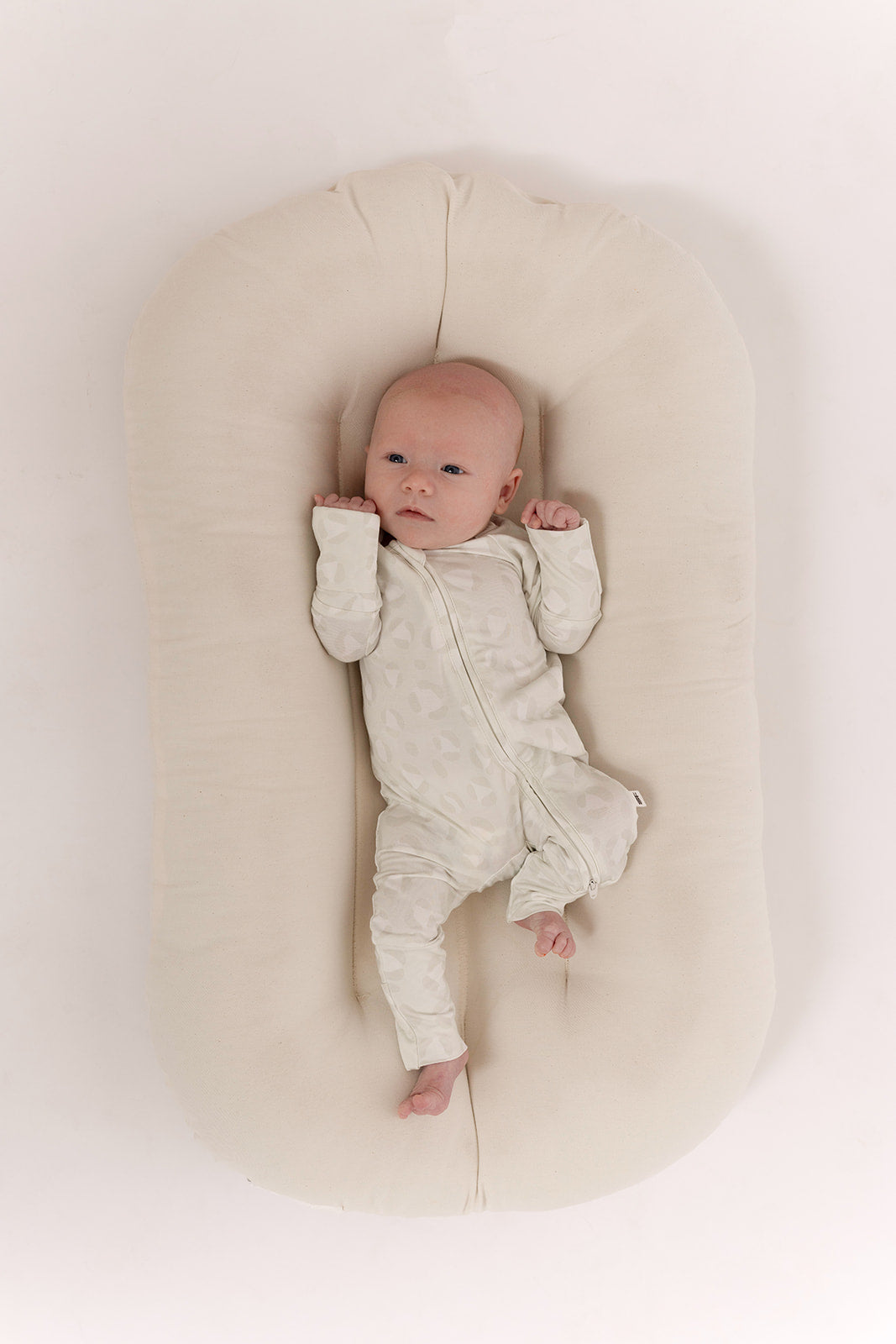A baby wearing forever french baby Bamboo Zip Pajamas | Meow rests on a soft, oval beige cushion with arms raised, gazing upward against a plain white background.