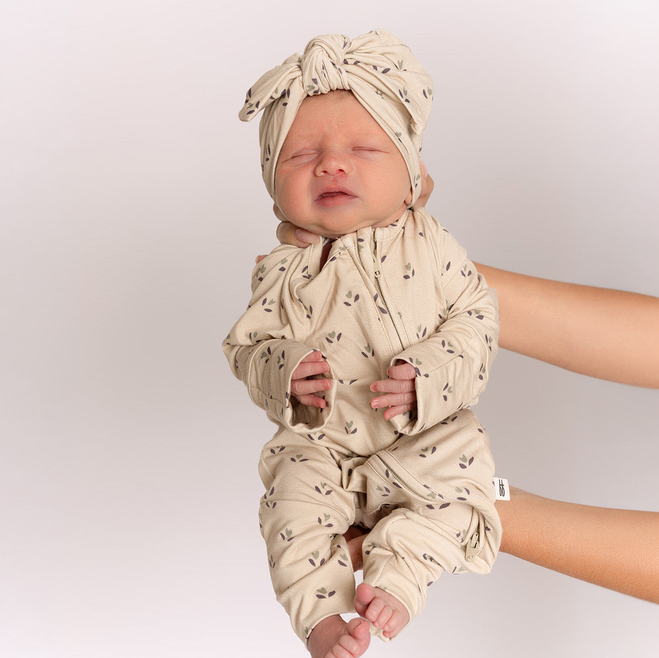 A newborn in forever french baby’s Bamboo Zip Pajamas | Flora—a beige patterned onesie with a matching head wrap—is gently held upright by an adult against a simple light background.
