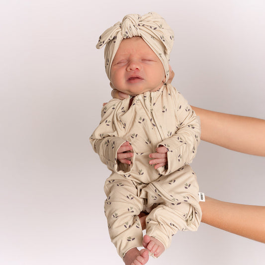 A newborn in forever french baby’s Bamboo Zip Pajamas | Flora—a beige patterned onesie with a matching head wrap—is gently held upright by an adult against a simple light background.