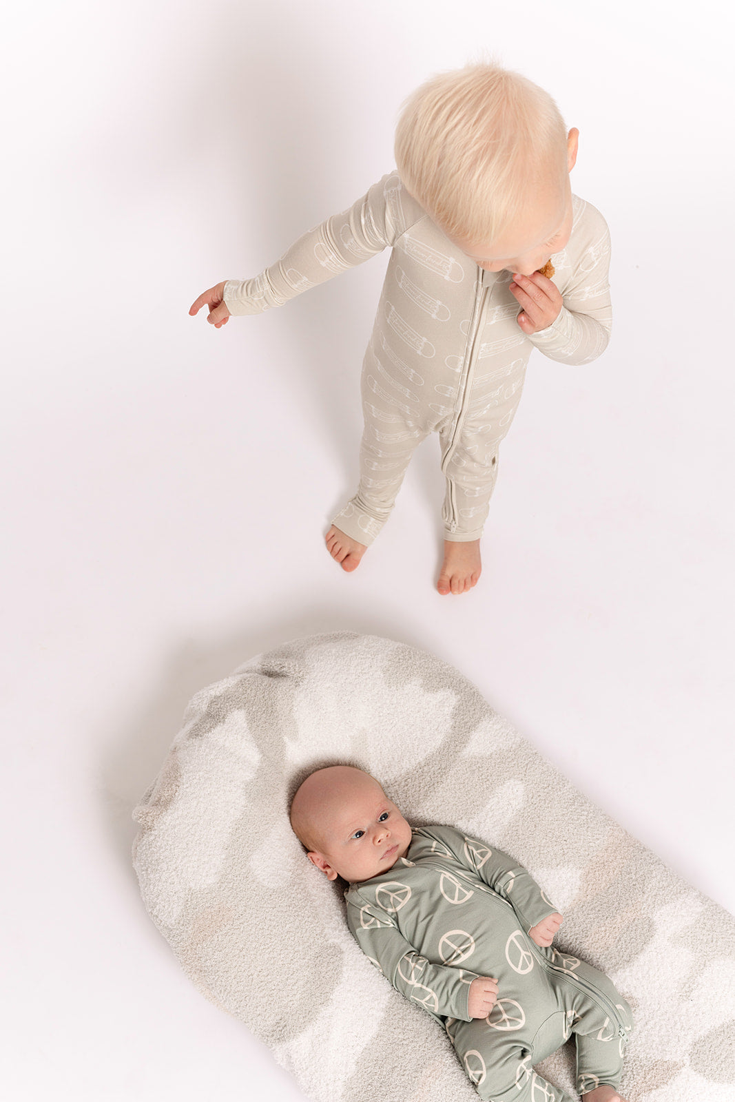 A baby in forever french baby's Bamboo Zip Pajamas | Peace lies on a soft, patterned blanket while an older toddler stands nearby, both in hypoallergenic, light-colored onesies designed for sensitive skin on a white background.