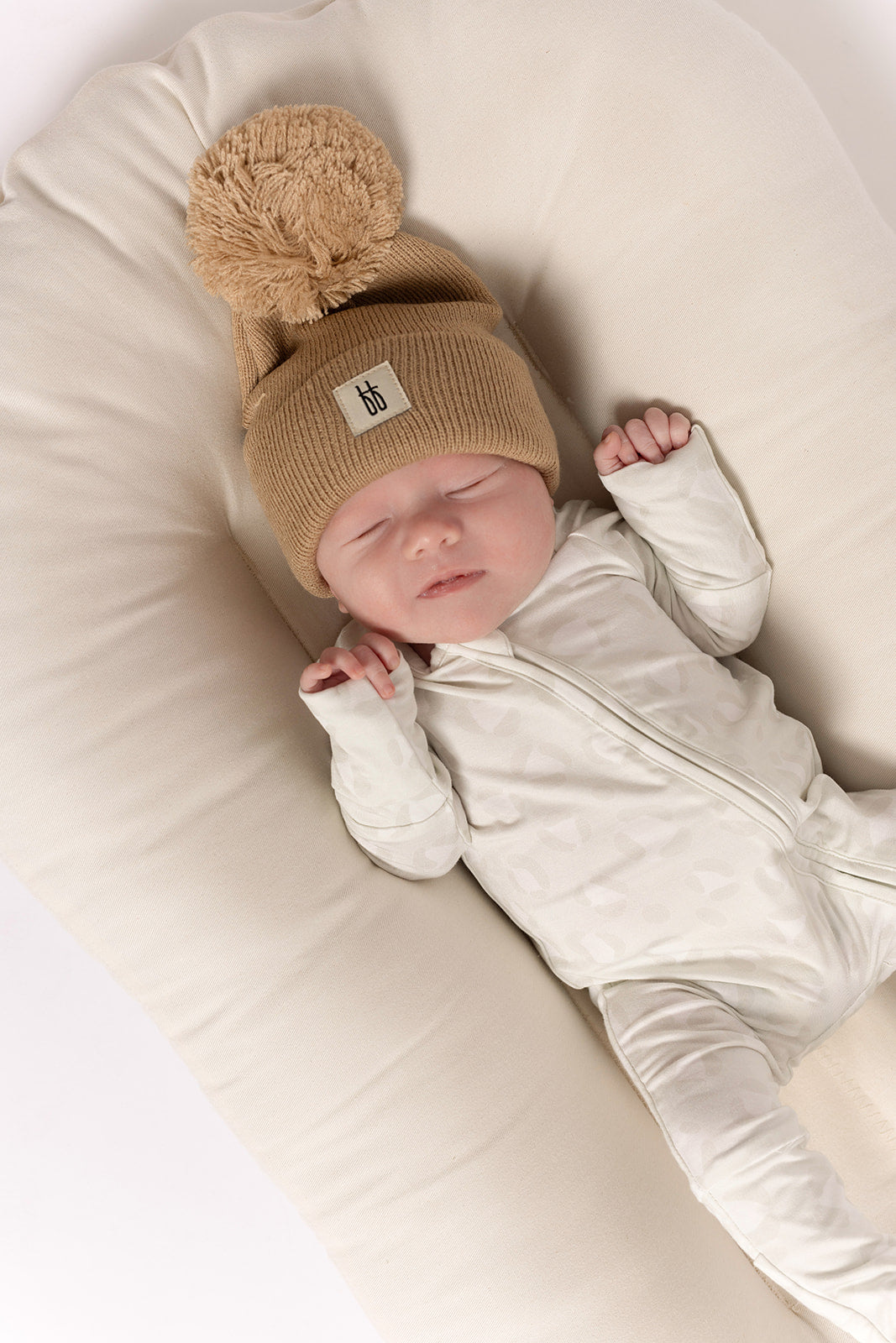 A newborn sleeps on a soft, cream cushion, wearing forever french baby’s Bamboo Zip Pajamas | Meow—a cozy, hypoallergenic white zip-up onesie—and a beige knit hat with pom-pom. Their eyes are closed and hands gently curled.