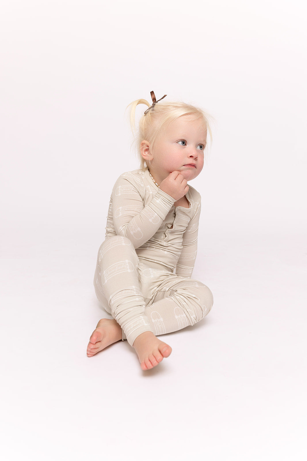 A young child with blonde pigtails sits barefoot on a white background, wearing forever french baby Bamboo Two Piece Pajamas | Sk8er Boy in light beige with a subtle pattern, gazing upward thoughtfully.