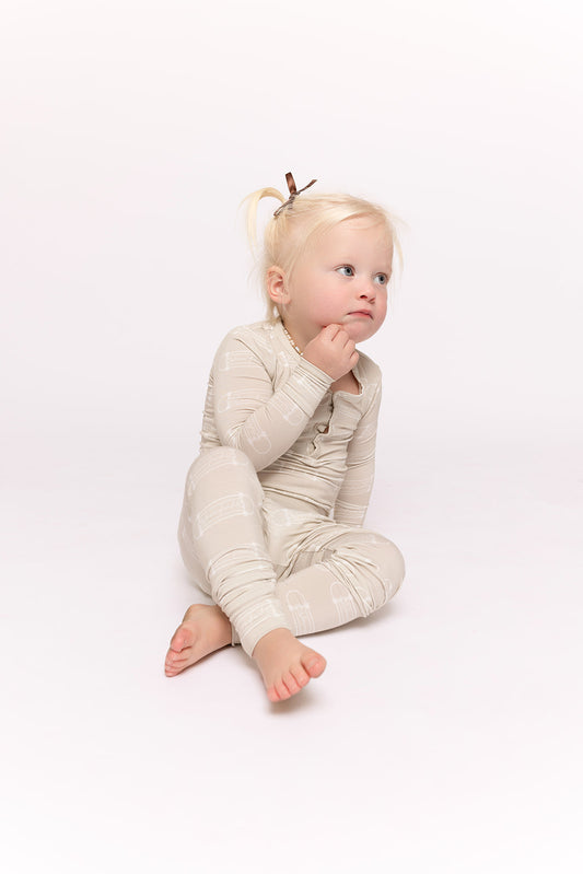 A young child with blonde pigtails sits barefoot on a white background, wearing forever french baby Bamboo Two Piece Pajamas | Sk8er Boy in light beige with a subtle pattern, gazing upward thoughtfully.