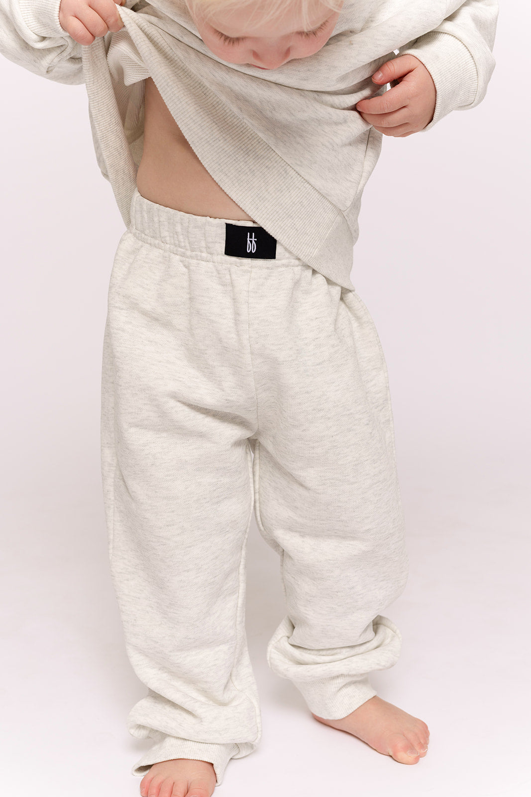 A young child wears the forever french baby Child Sweat Set in Heather Grey, standing barefoot against a light background—a cozy and stylish choice for everyday kids' wear.