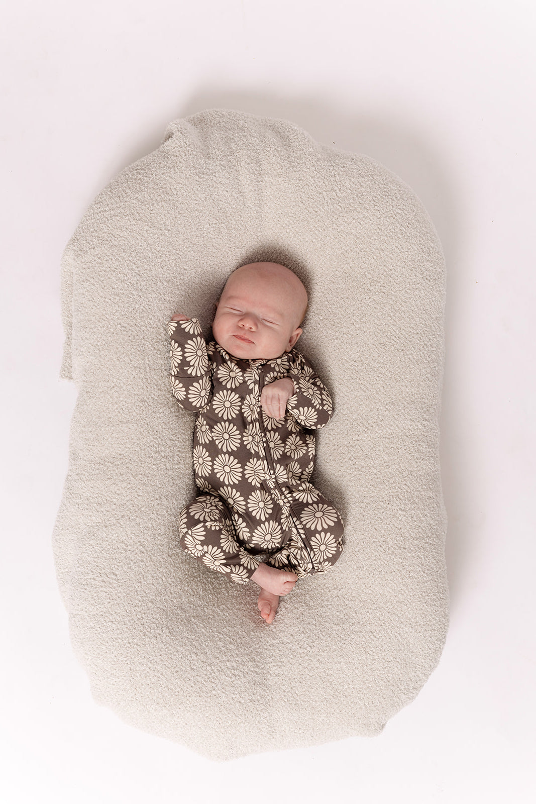 A baby sleeps in forever french baby Bamboo Zip Pajamas | Retro Floral—a brown, daisy-patterned onesie—on a soft light beige blanket, photographed from above on a white background.