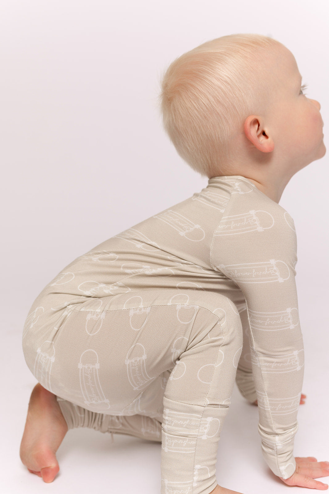 A young child with short blond hair is squatting on the floor, wearing forever french baby's Bamboo Zip Pajamas | Sk8er Boy—a light beige, long-sleeve bamboo set with a subtle white pattern—against a plain background.