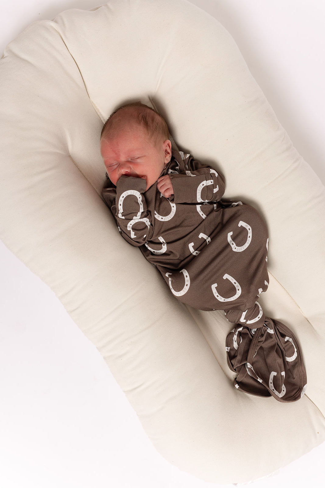 A newborn sleeps on a soft off-white cushion, wrapped in the forever french baby Bamboo Swaddle | Giddy Up featuring a brown background with white horseshoes, eyes closed and one hand by their face.