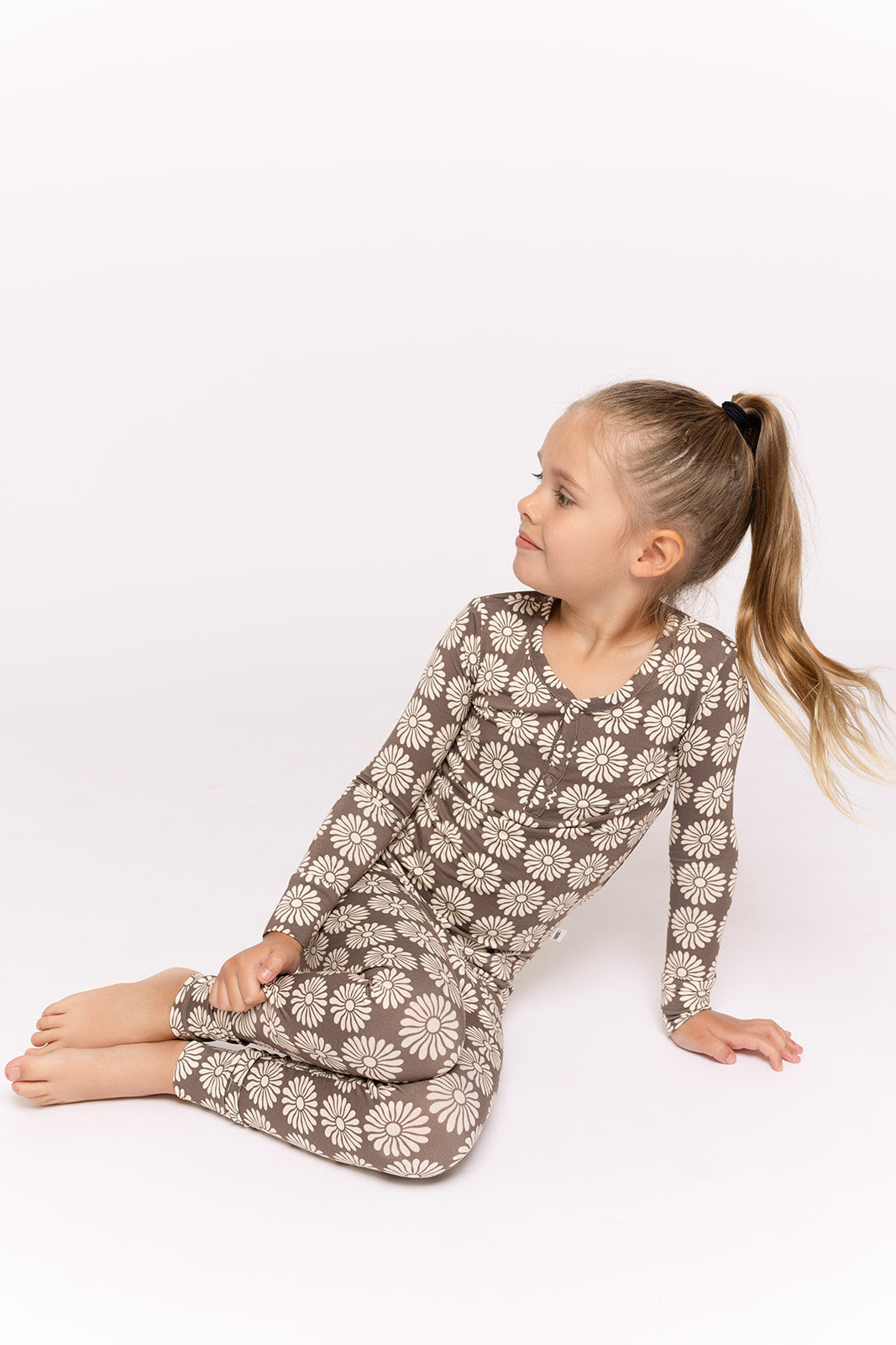 A young girl with long blonde hair in a ponytail sits barefoot on the floor, wearing forever french baby Bamboo Two Piece Pajamas in Retro Floral, looking to her left against a plain white background.