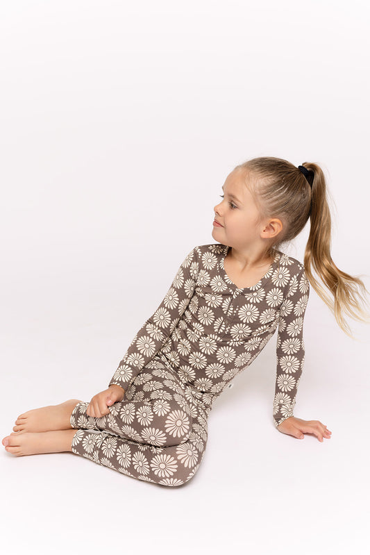 A young girl with long blonde hair in a ponytail sits barefoot on the floor, wearing forever french baby Bamboo Two Piece Pajamas in Retro Floral, looking to her left against a plain white background.